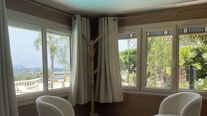 A room with two white chairs, a round table, and a woven stool. There are large windows with curtains, a view of greenery and buildings outside. - Hôtel Miramar (Vence)