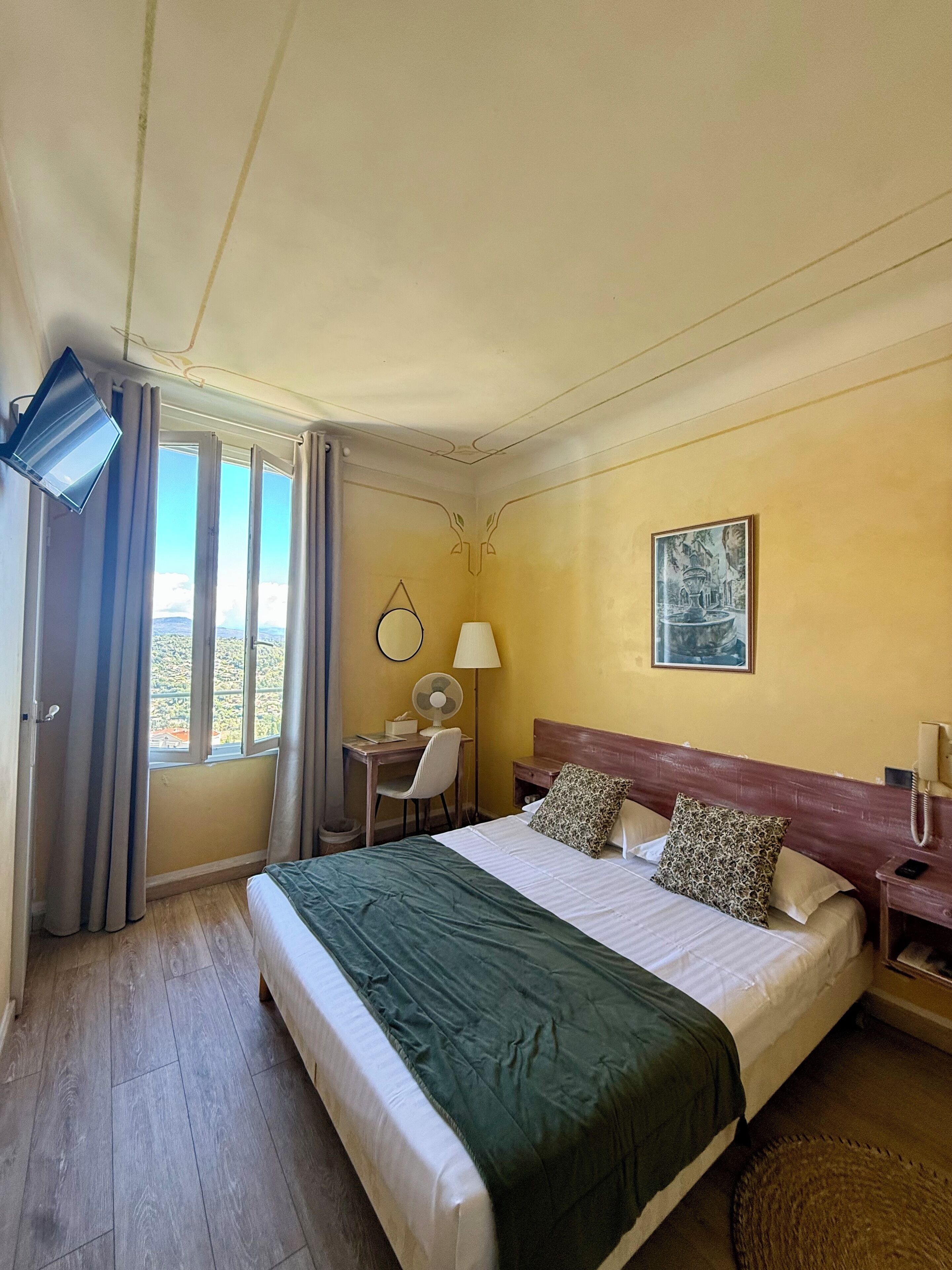 A hotel room with a bed, a desk, a chair, a lamp, a telephone, and a painting on the wall.
