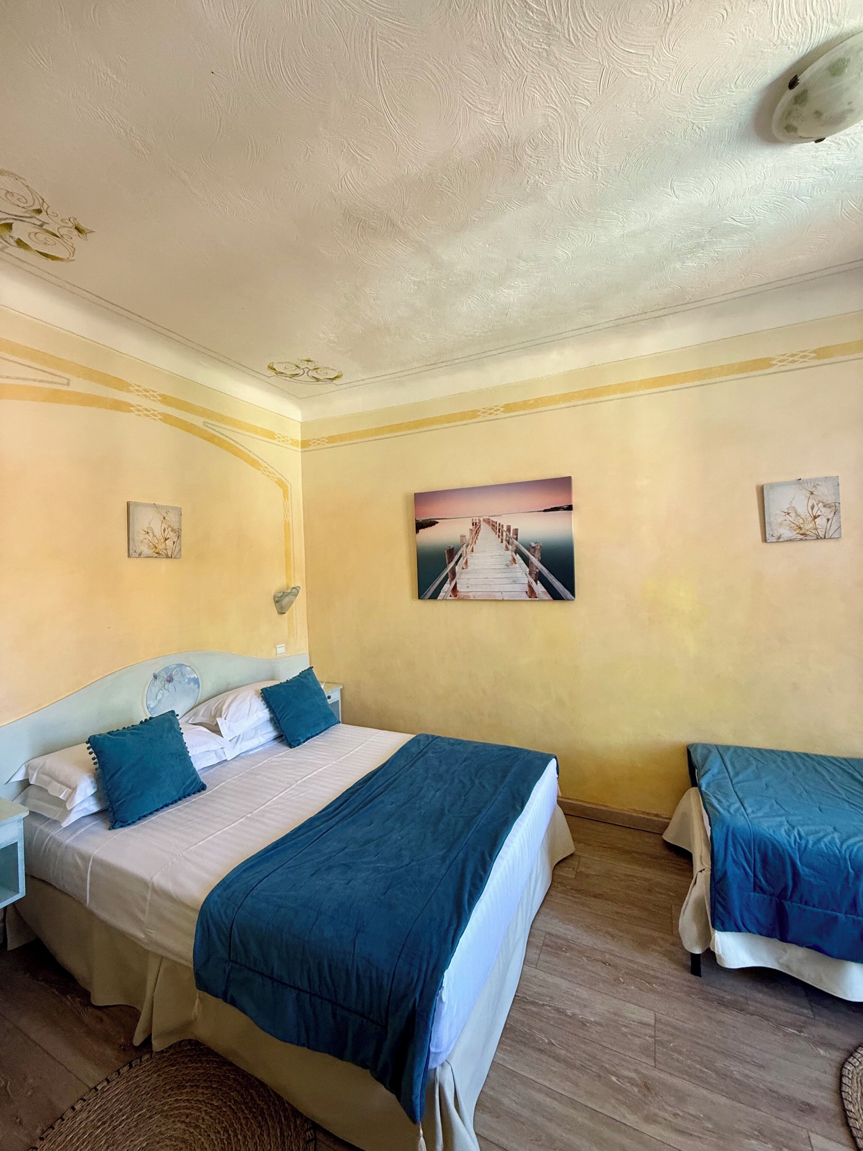 A hotel room with two beds, a large painting of a pier, and a small table.