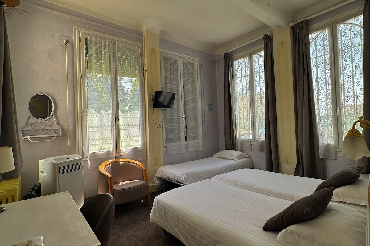 A hotel room with two beds, a desk, a chair, a TV, and large windows with curtains.