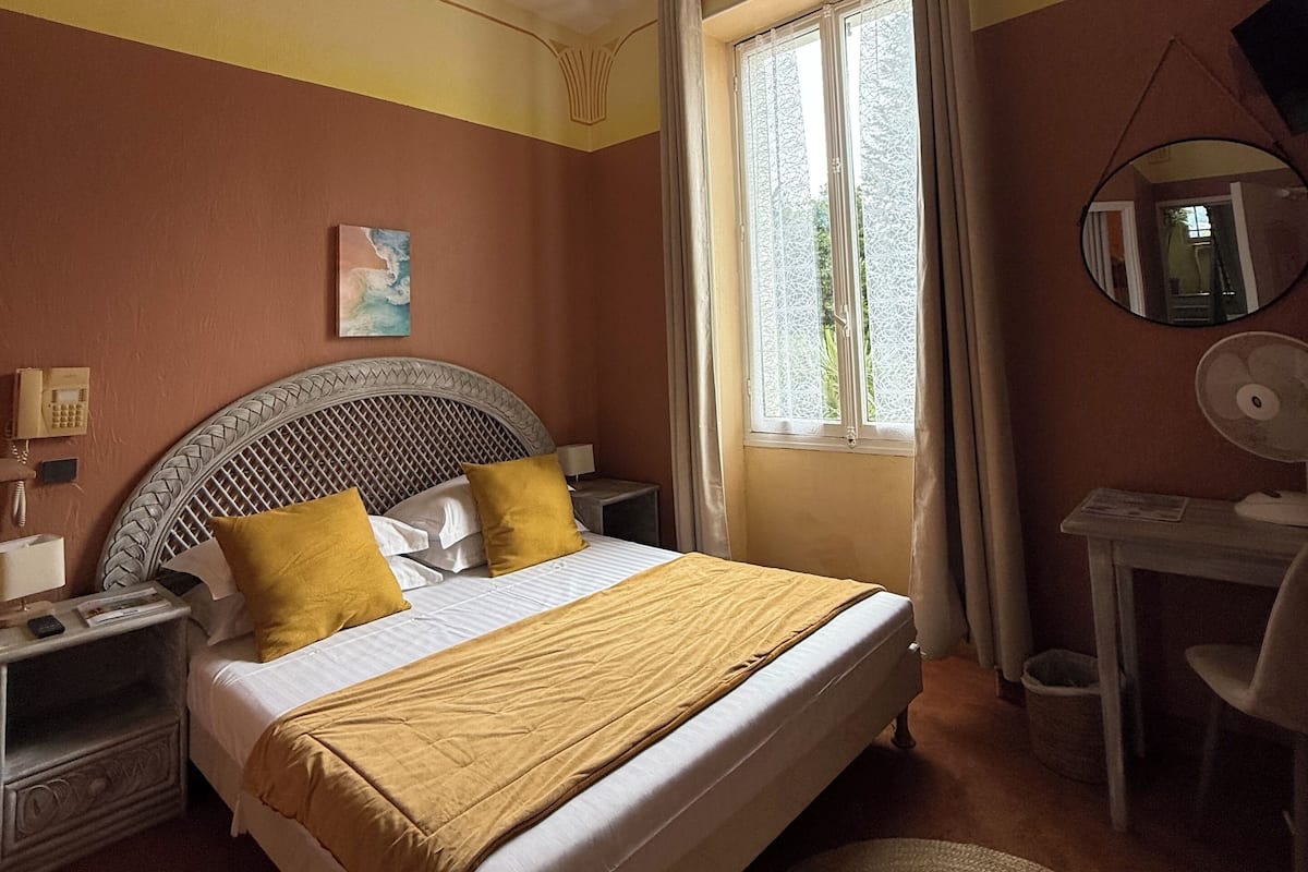 A hotel room with a bed, a desk, a chair, a bedside table, a telephone, and a window with curtains.