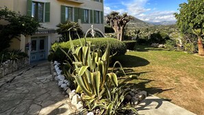 A hotel with a stone pathway leading to the entrance, a well-maintained garden with various plants, and a clear view of the surrounding landscape. - Hôtel Miramar (Vence)