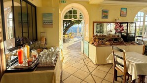 A hotel breakfast area with a buffet, a view of the sea through a large window, and a dining area with a table and chairs. - Hôtel Miramar (Vence)