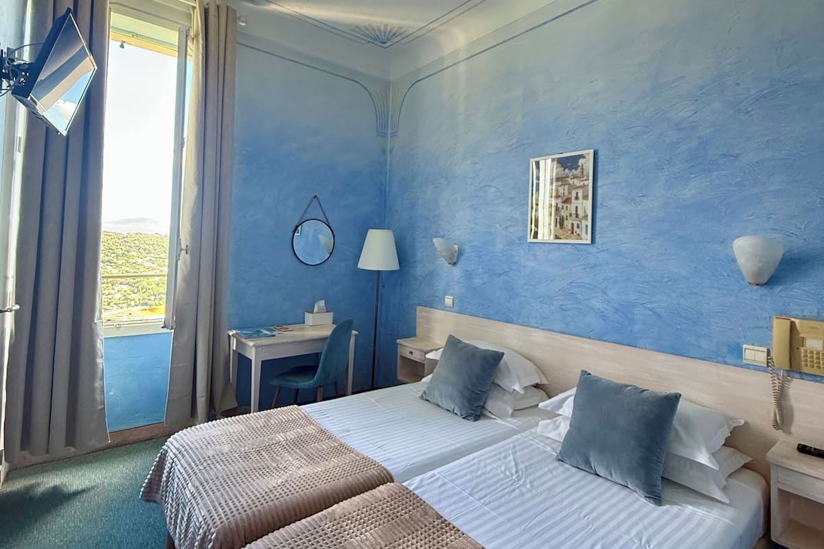 A hotel room with two beds, a desk, a chair, a lamp, and a view of the outdoors.