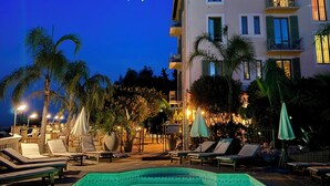 A swimming pool with lounge chairs and umbrellas, surrounded by palm trees and a building with multiple balconies. - Hôtel Miramar (Vence)