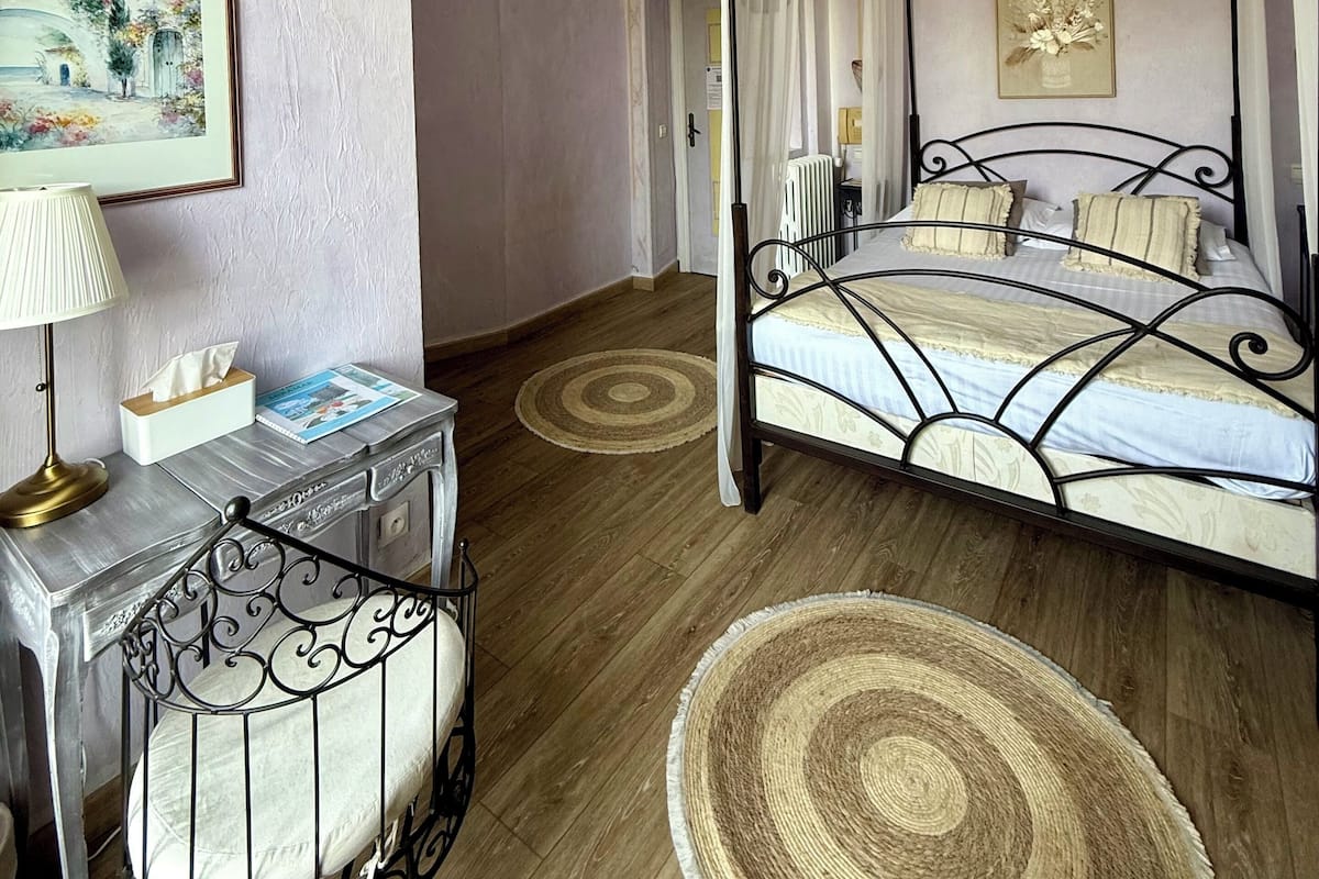 A four-poster bed with a canopy, a bedside table with a lamp, a framed painting on the wall, and a round rug on the floor.