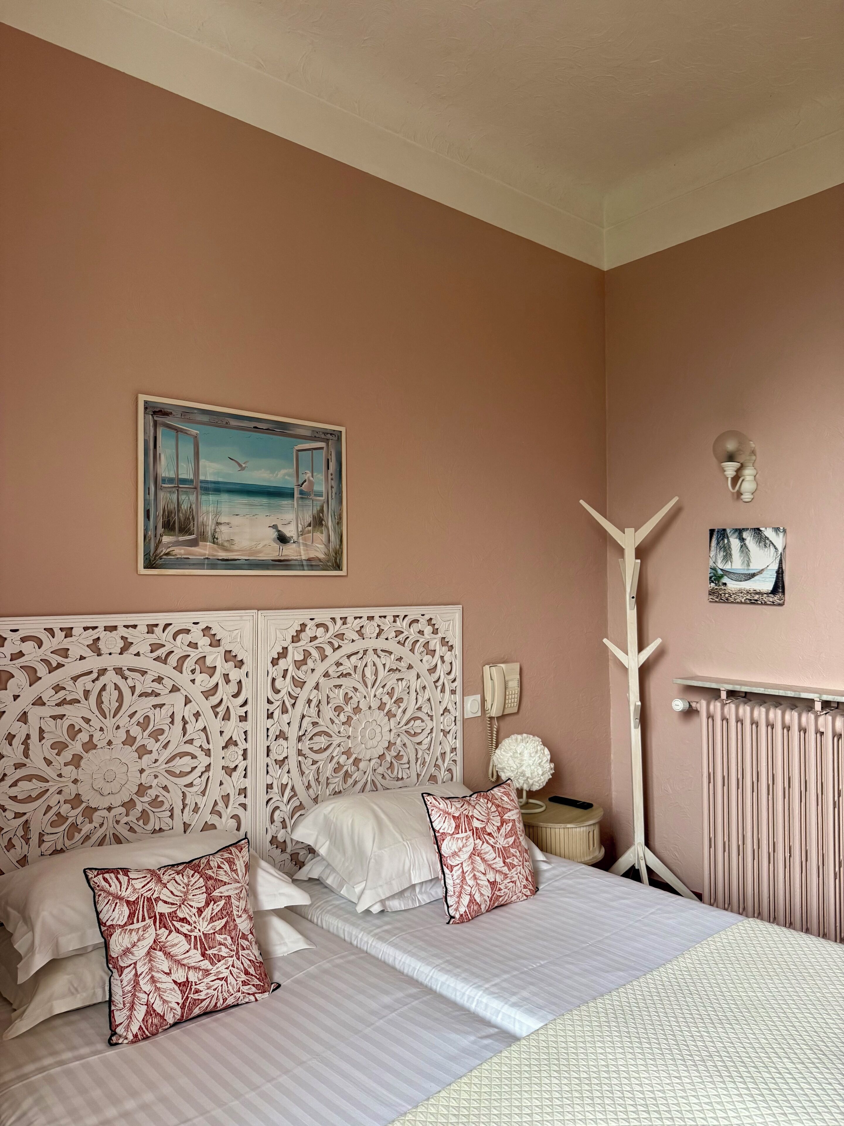 A bedroom with a large bed, a decorative headboard, a painting of a beach scene, a radiator, and a wall-mounted telephone.