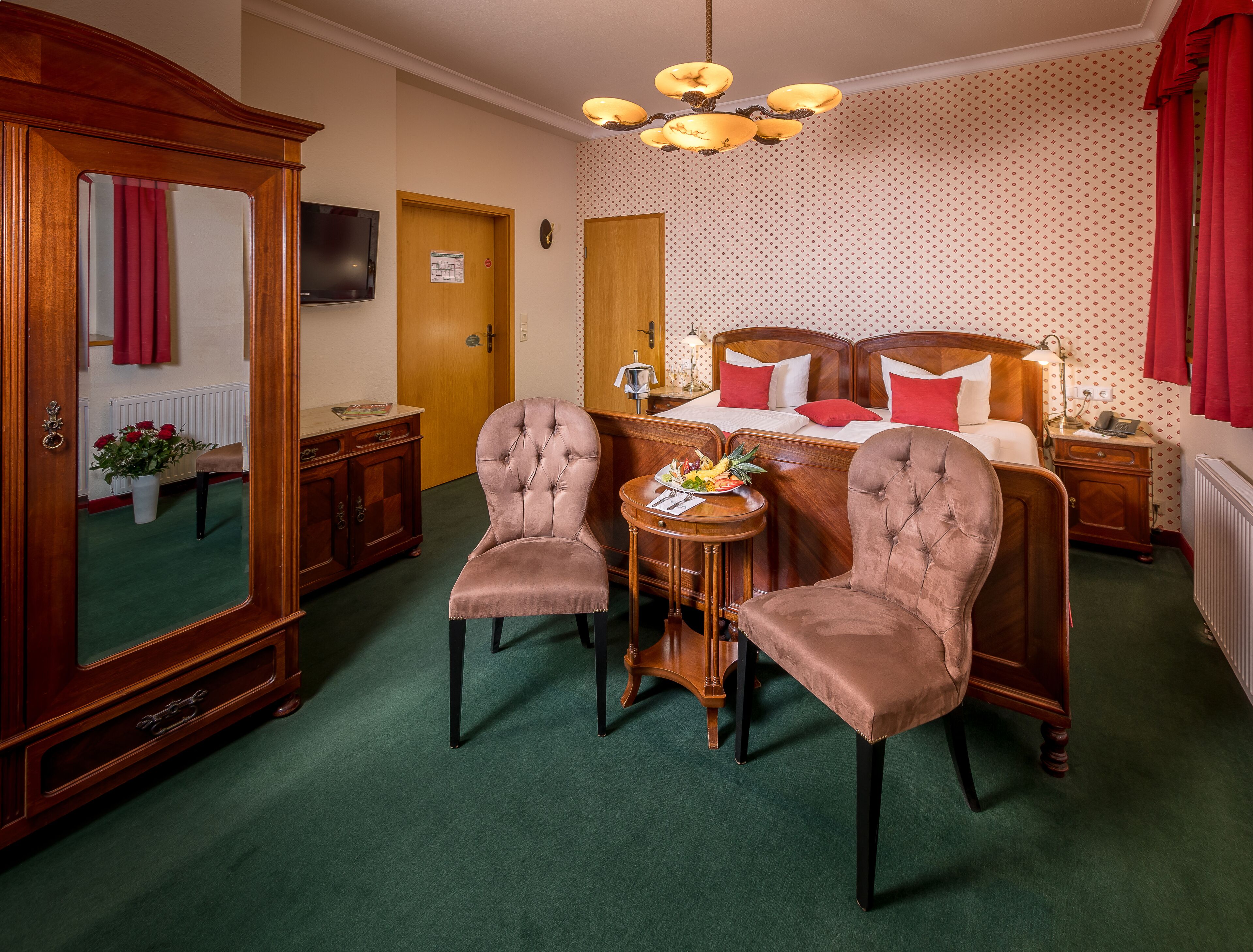 A hotel room with a large mirror, two upholstered chairs, a wooden bed with red pillows, a small table with fruit, and a chandelier.