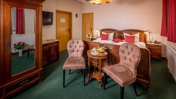 A hotel room with a large mirror, two upholstered chairs, a wooden bed with red pillows, a small table with fruit, and a chandelier.