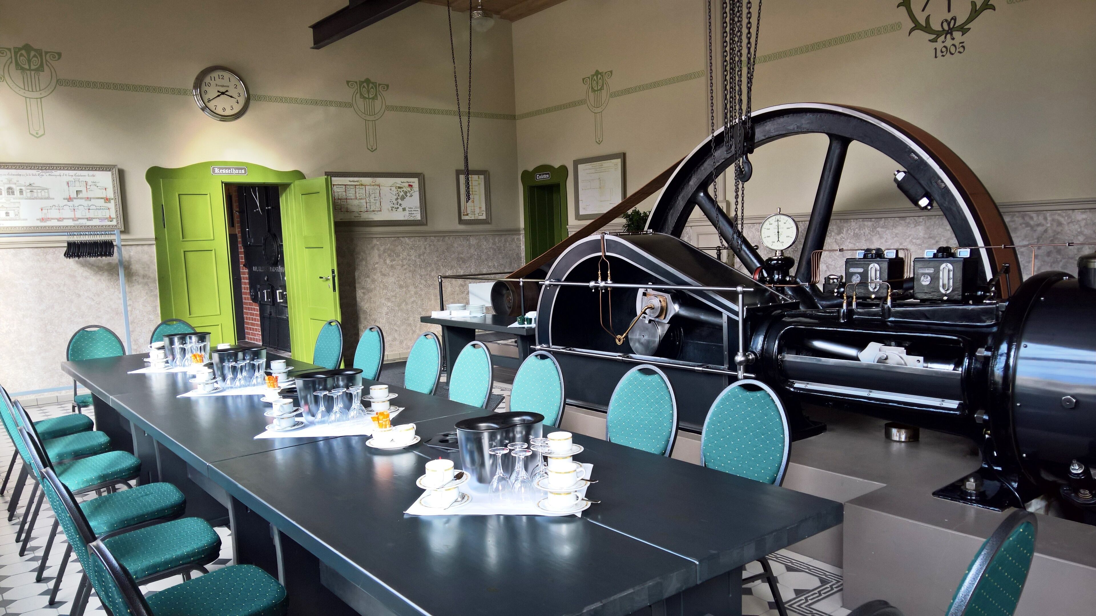 Un comedor con una larga mesa dispuesta para una comida, sillas verdes y una locomotora de vapor antigua al fondo.