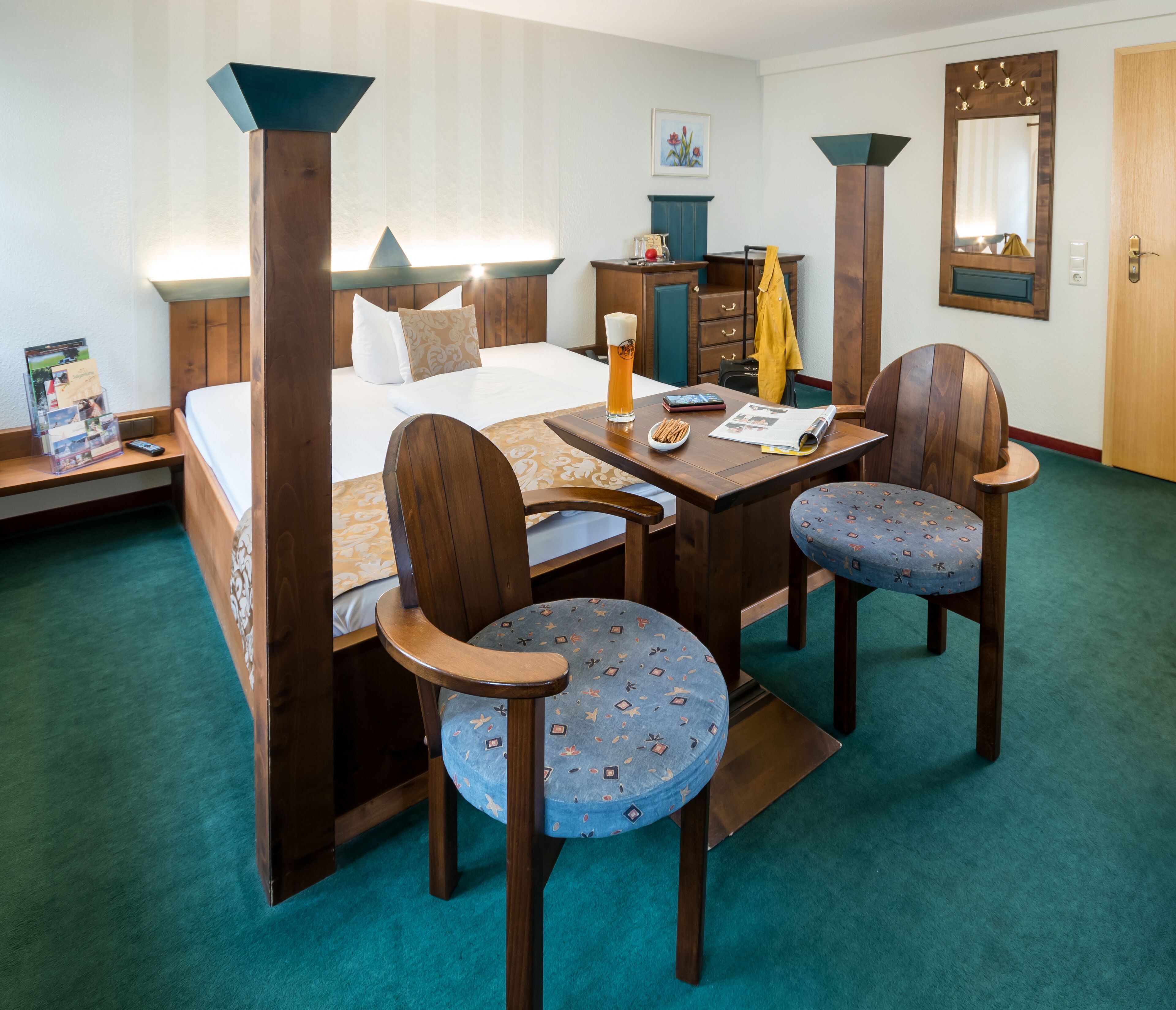 A hotel room with a bed, a desk with a chair, a mirror, and a wooden wardrobe.