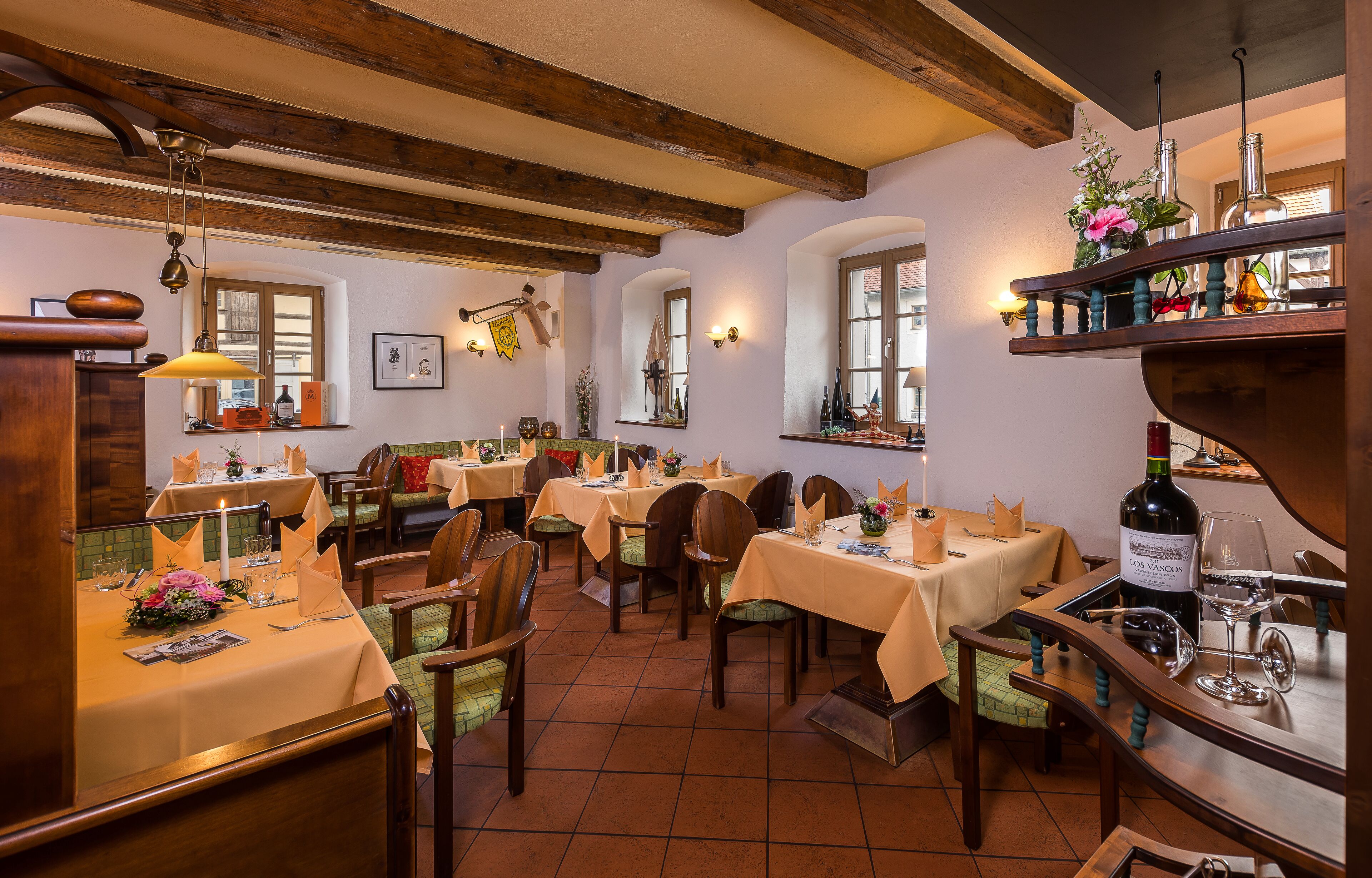 A restaurant with wooden beams, tables set for dining, and wine bottles on the bar.