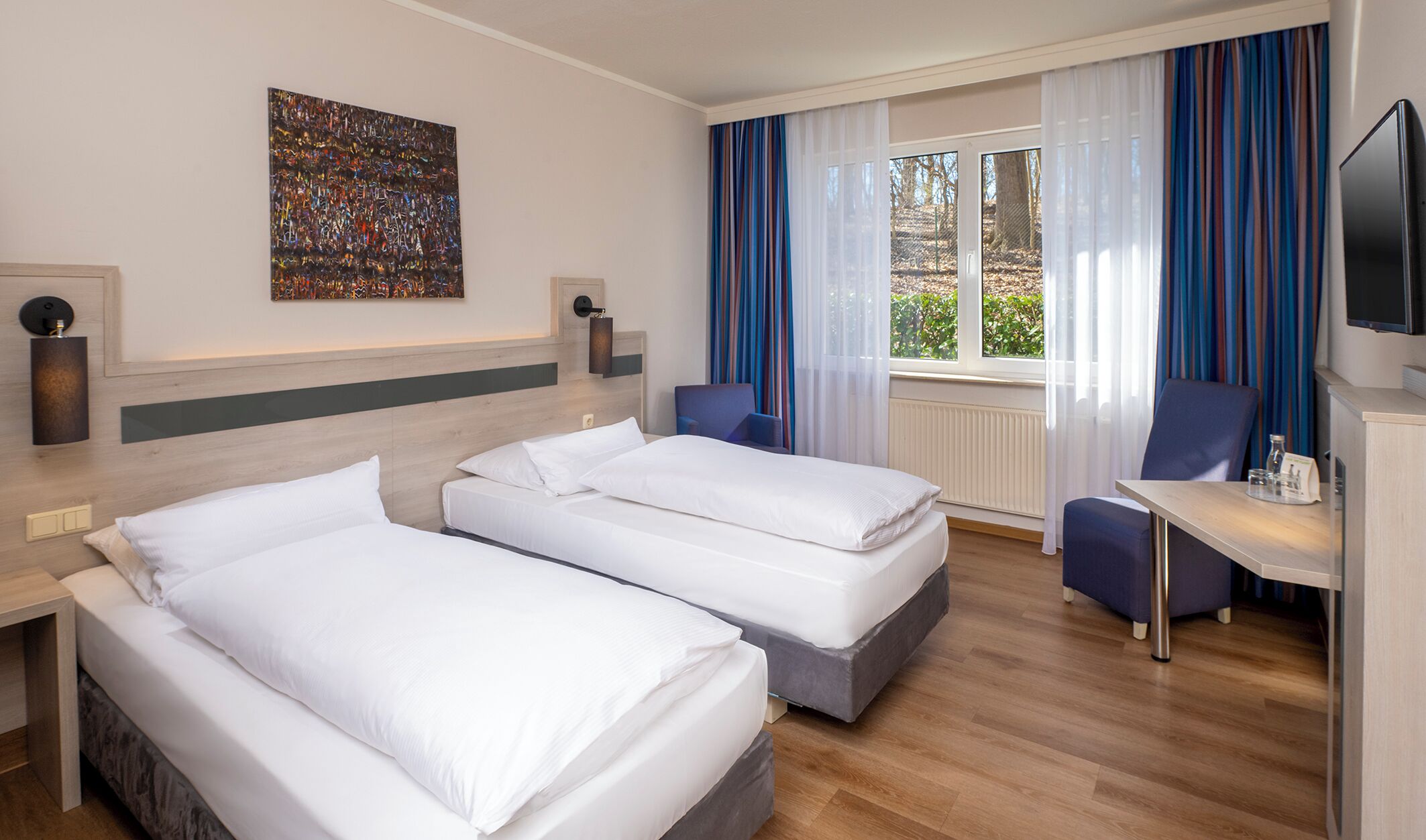A hotel room with two beds, a desk, a chair, a TV, and a window with curtains.