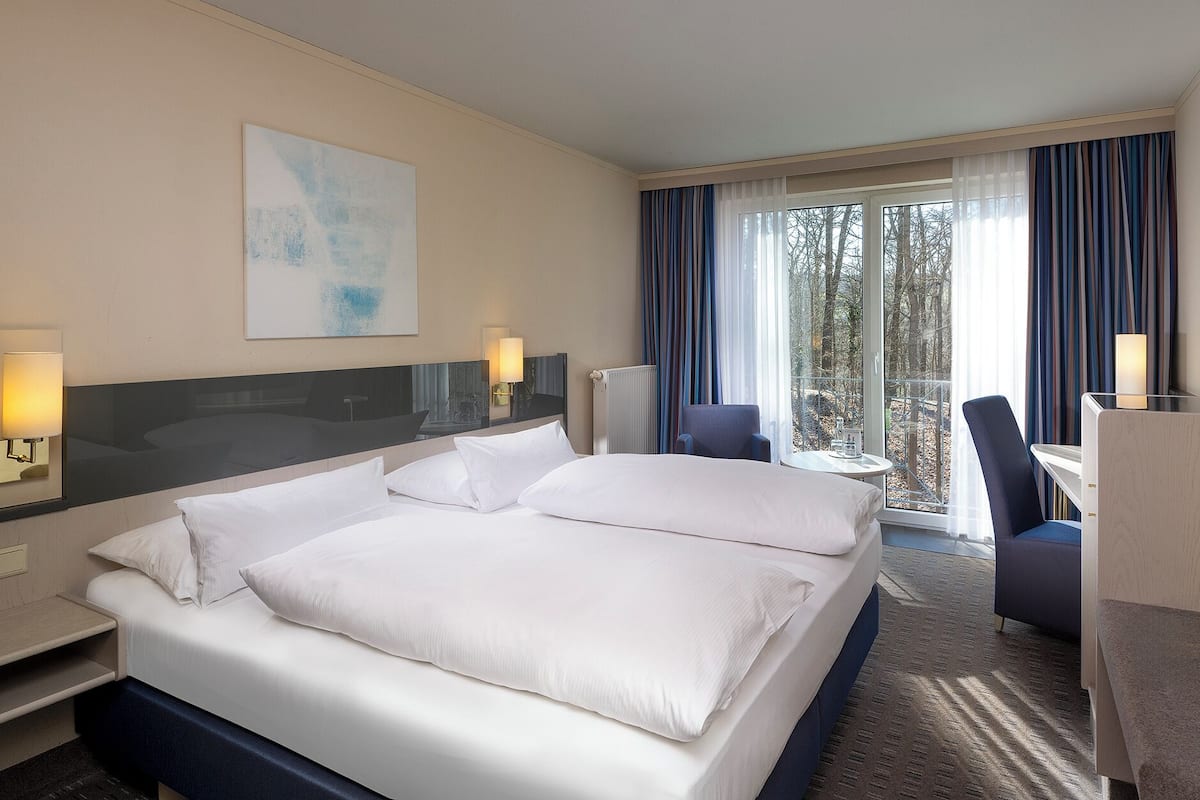 Ein Hotelzimmer mit einem großen Bett, einem Schreibtisch, einem Sessel, einem Fernseher und Blick ins Freie.