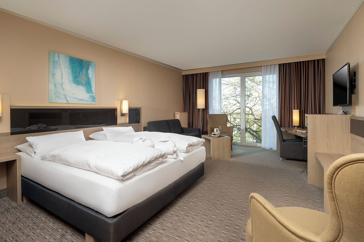 Ein Hotelzimmer mit einem großen Bett, einem Schreibtisch, einem Stuhl, einem Fernseher und einem Fenster mit Vorhängen.