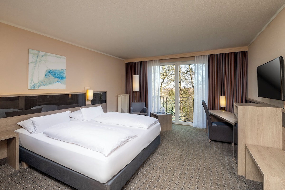 Ein Hotelzimmer mit einem großen Bett, einem Schreibtisch, einem Sessel, einem Fernseher und einem Fenster mit Vorhängen.