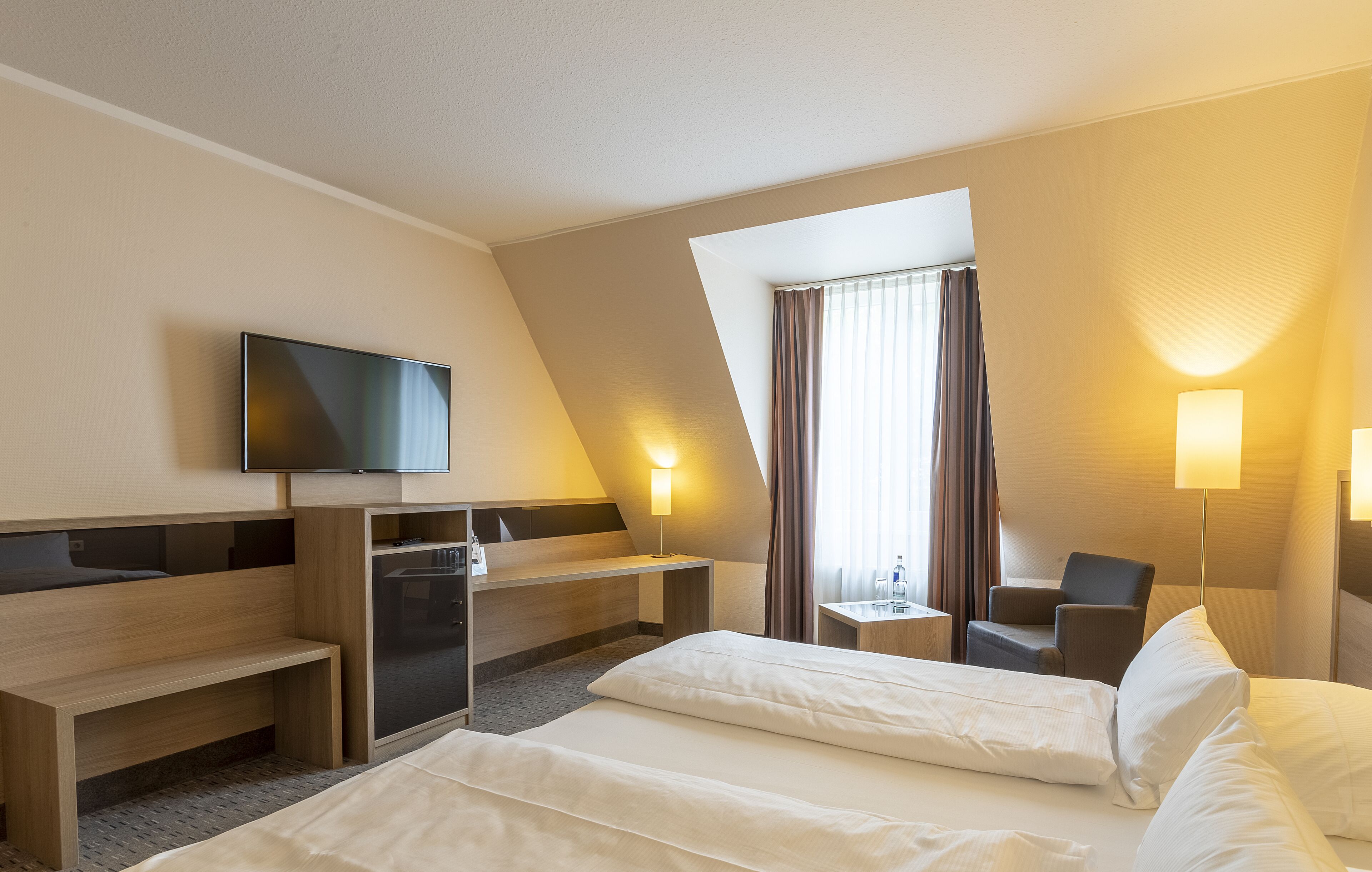 Una habitación de hotel moderna con cama, escritorio, silla, televisor y ventana con cortinas.