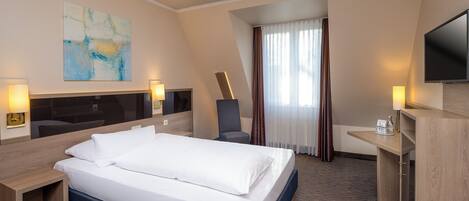 A hotel room with a bed, desk, chair, TV, and artwork on the wall.