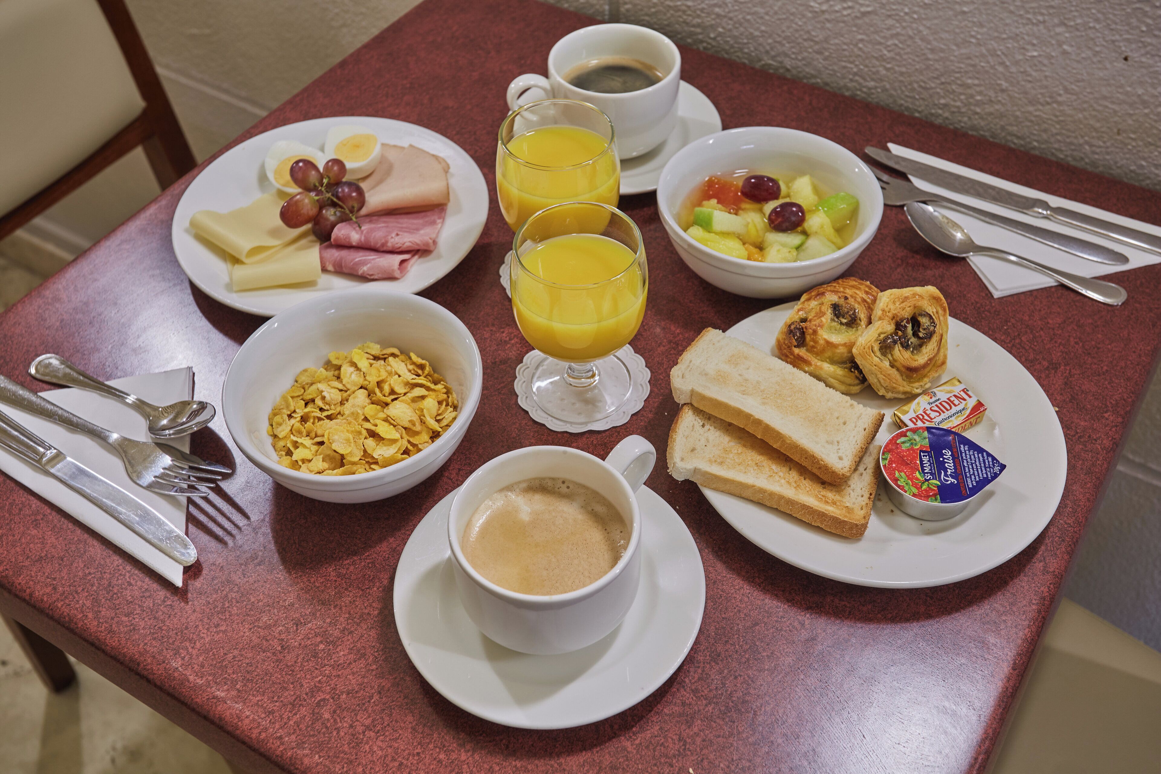 A breakfast table with plates of eggs, ham, cheese, toast, cereal, coffee, juice, and a bowl of fruit.