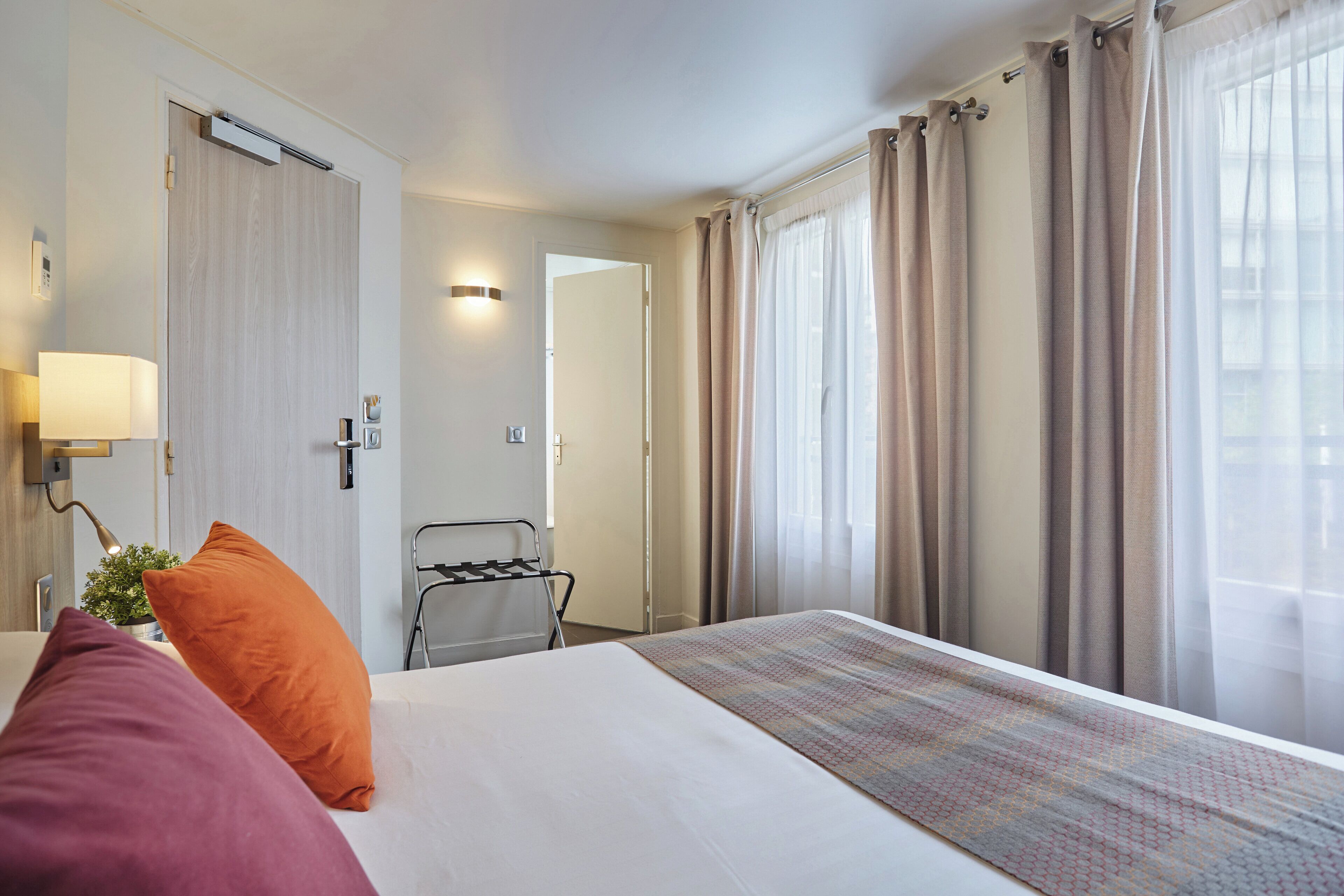 A hotel room with a bed, pillows, a lamp, a plant, and a window with curtains.