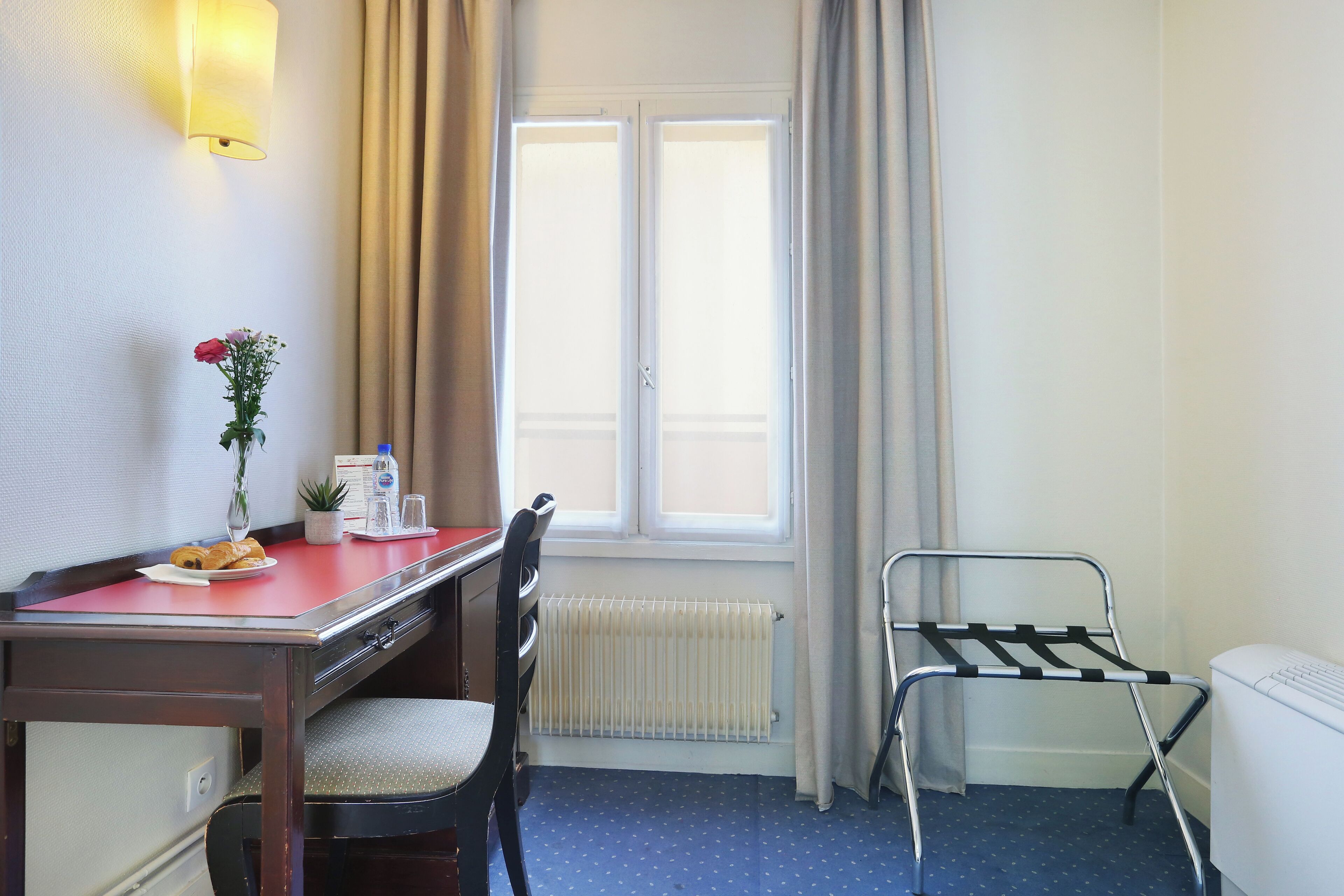 Ein Hotelzimmer mit Bett, Schreibtisch mit Stuhl, Heizkörper, Fenster mit Vorhängen und einem kleinen Tisch mit Vase und einer Schüssel Essen.