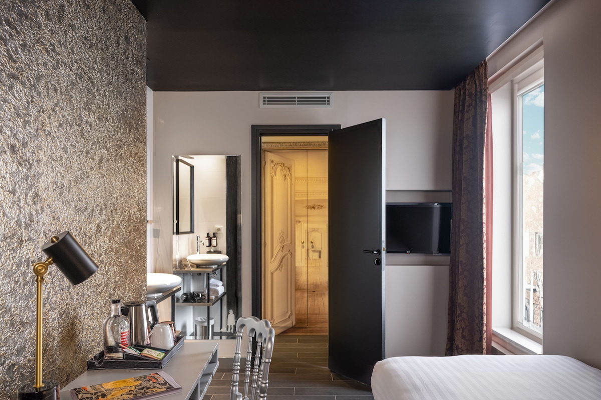 A modern hotel room with a bed, a desk with a lamp, a TV, and a bathroom visible through an open door.