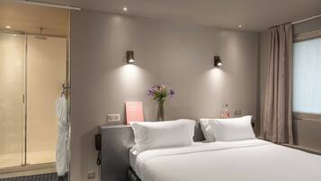 A hotel room with a bed, a shower area, a bedside table with a phone, a vase with flowers, and a sign on the wall.