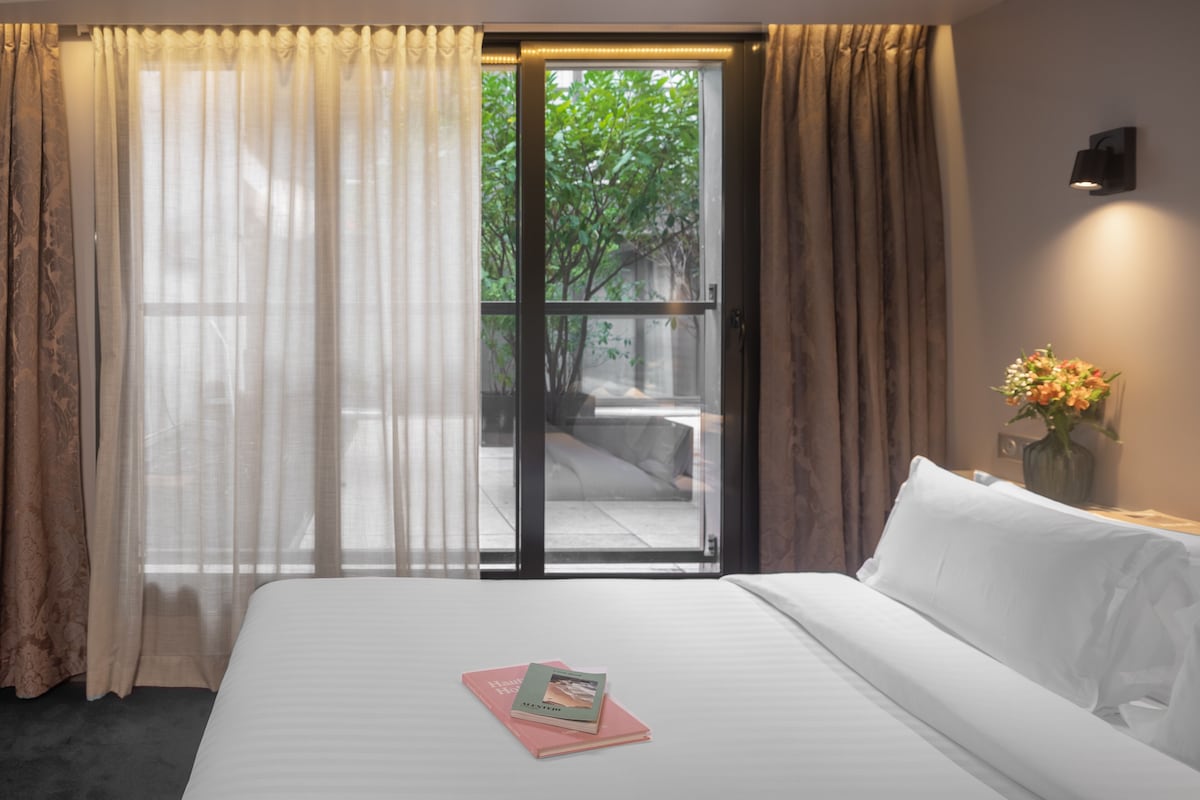 A hotel room with a bed, a window with curtains, a view of greenery outside, and a vase with flowers on the nightstand.