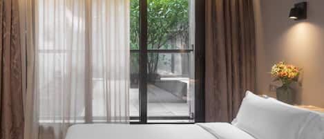 A hotel room with a bed, a window with curtains, a view of greenery outside, and a vase with flowers on the nightstand.
