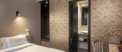 A modern hotel room with a large bed, bedside table, and a chair. There is a bathroom visible through an open door.