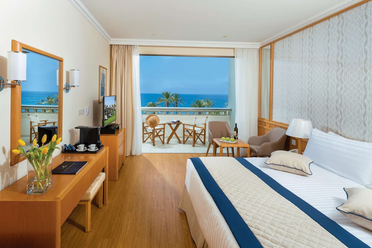 Une chambre d’hôtel avec un grand lit, un bureau avec un ordinateur, une chaise, une petite table avec une lampe, et une vue sur l’océan depuis un balcon.
