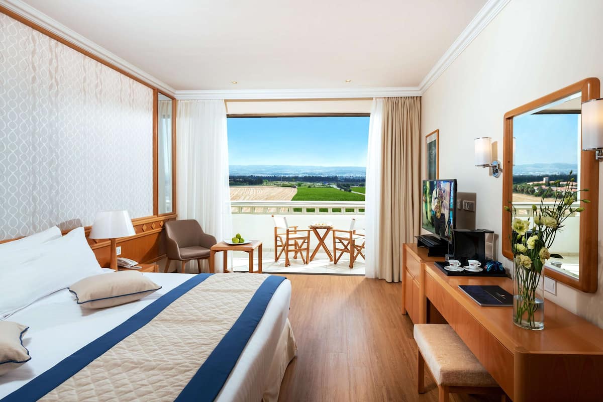 Une chambre d’hôtel avec un grand lit, un bureau avec un ordinateur, une télévision et un balcon offrant une vue sur la campagne.