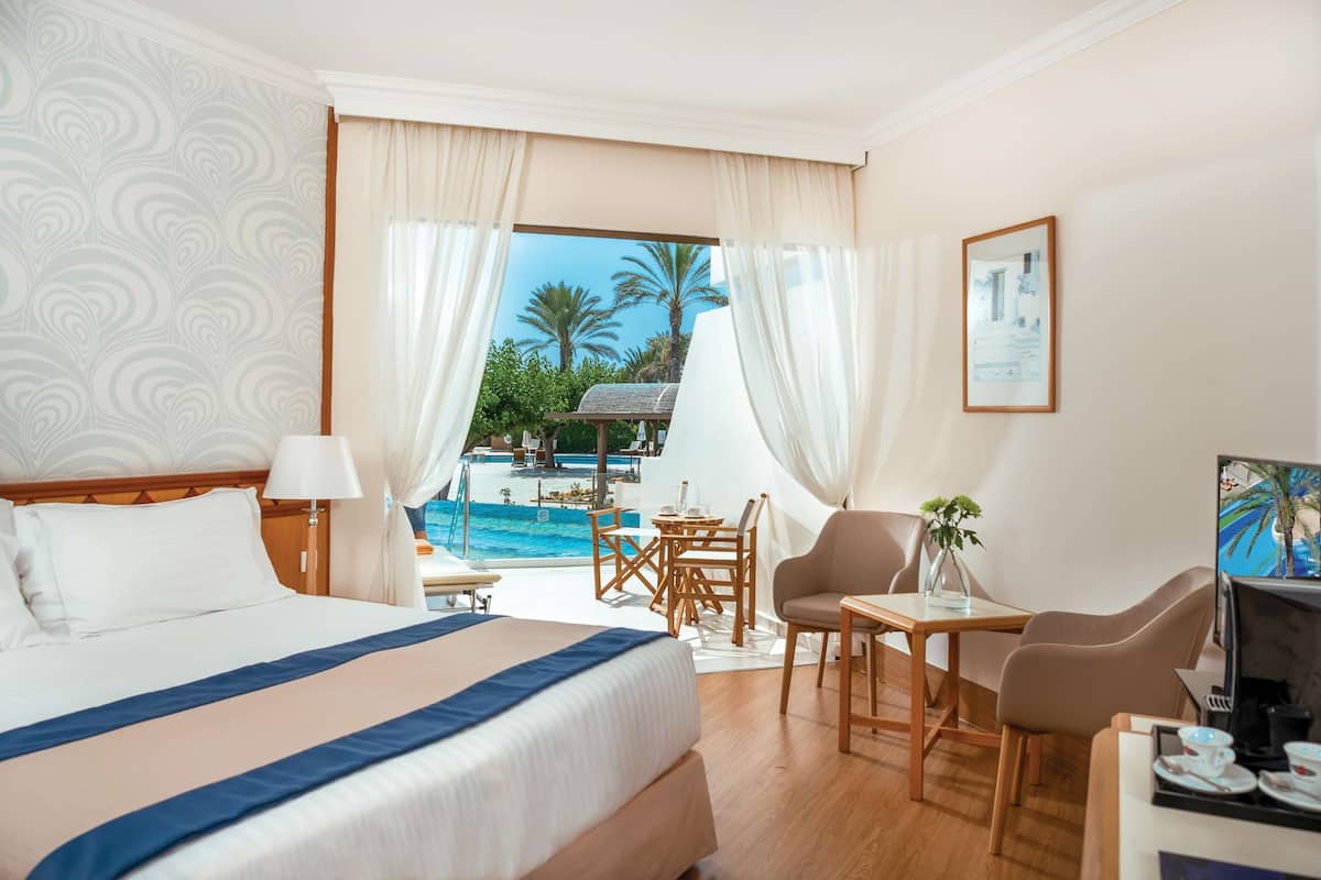 Une chambre d’hôtel avec un lit, un bureau, une chaise, une petite table et une vue sur une piscine et des palmiers.