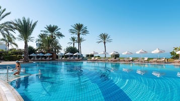 Piscina con sillas de descanso y sombrillas, rodeada de palmeras y un cielo despejado.