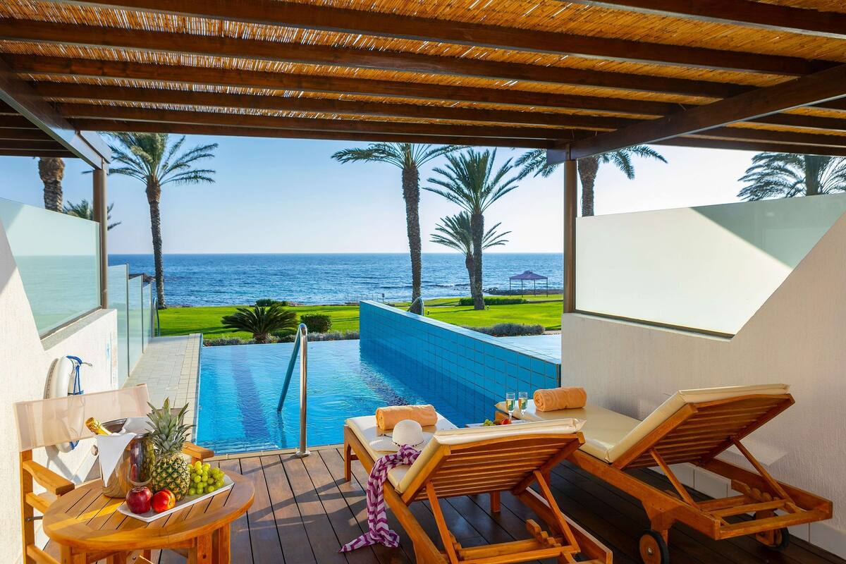 Un espace aménagé au bord de la piscine, avec des chaises longues, une table garnie de fruits et une vue sur la mer et les palmiers.