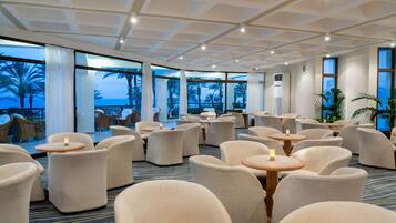 A hotel lobby with round tables and chairs, large windows with views of palm trees, and a ceiling with recessed lighting.