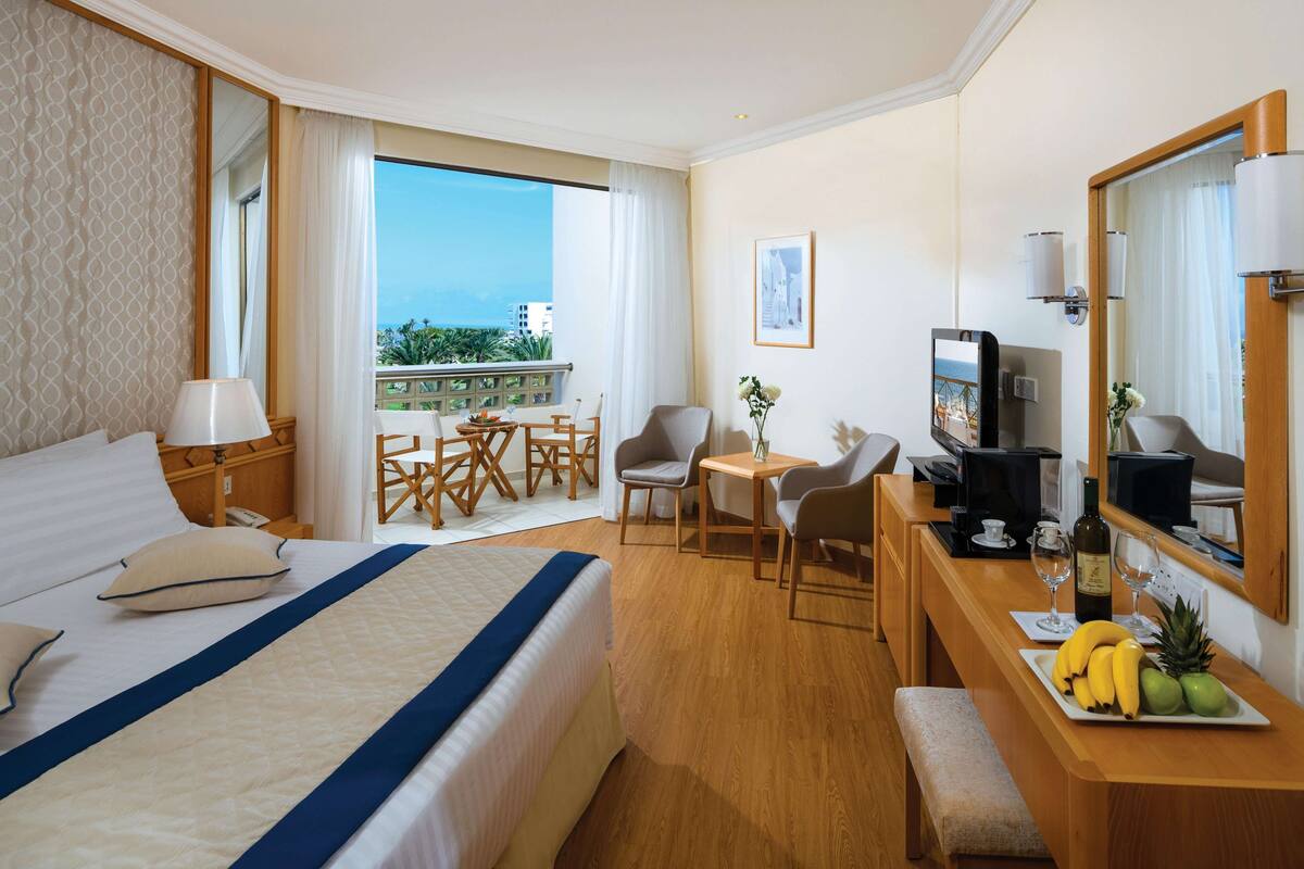 Une chambre d’hôtel avec un lit, un bureau sur lequel se trouve une assiette de fruits, une télévision et un balcon offrant une vue sur la ville.