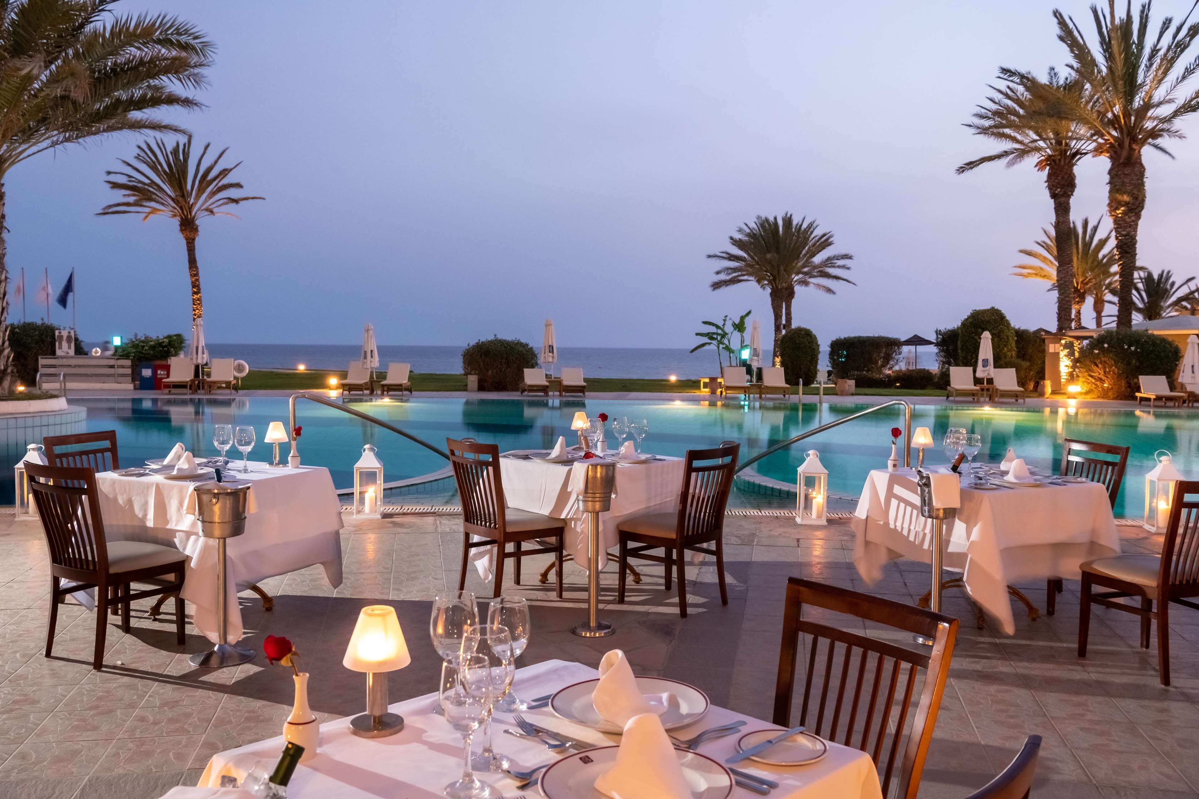 A poolside dining area with tables set for two, overlooking a pool and the sea.
