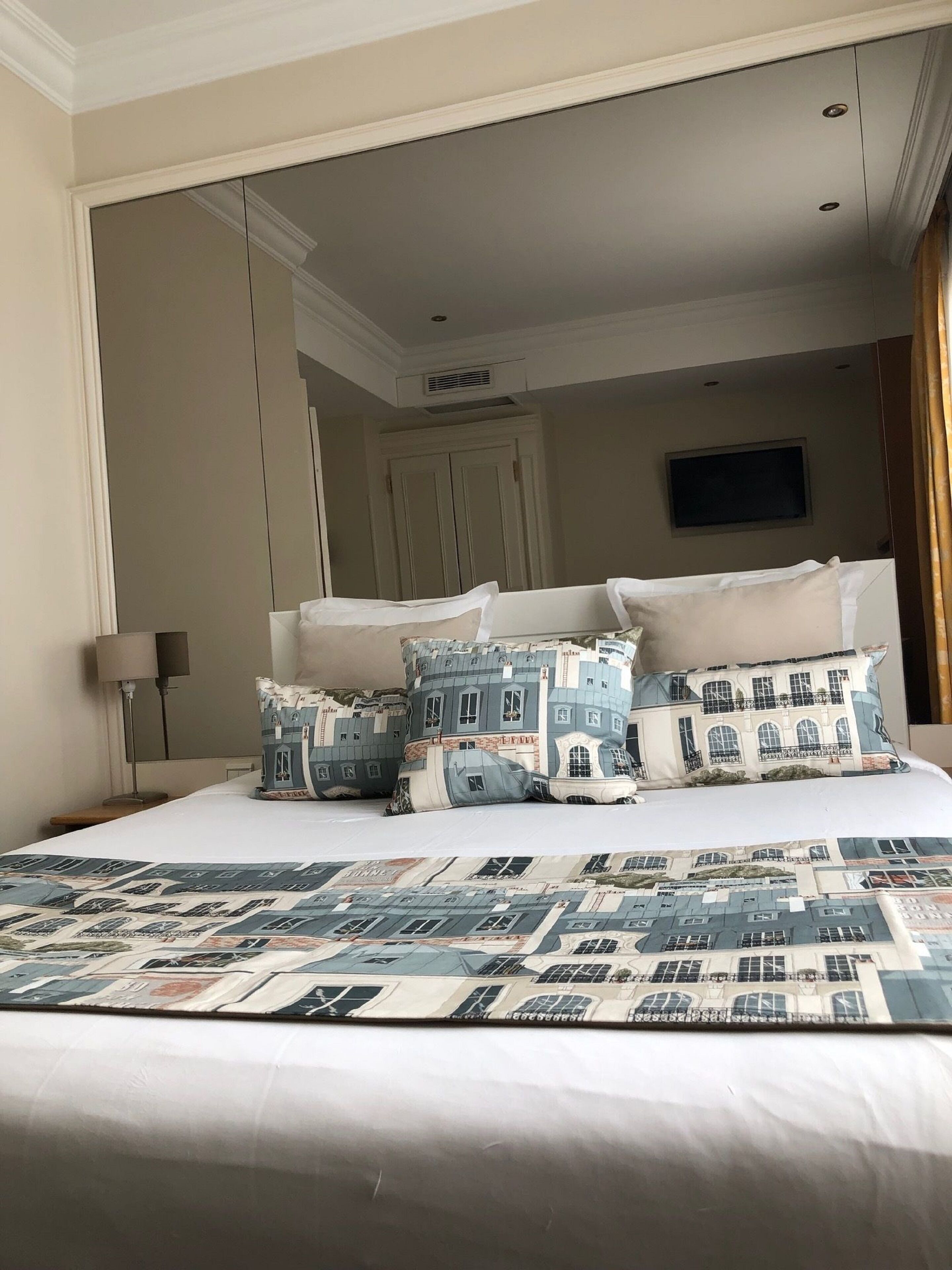 Una habitació d'hotel amb un llit gran, vàries almoines amb un dibuix d'edifici, un televisor pantalla plana muntat a la paret i una taula de nit amb una làmpada.