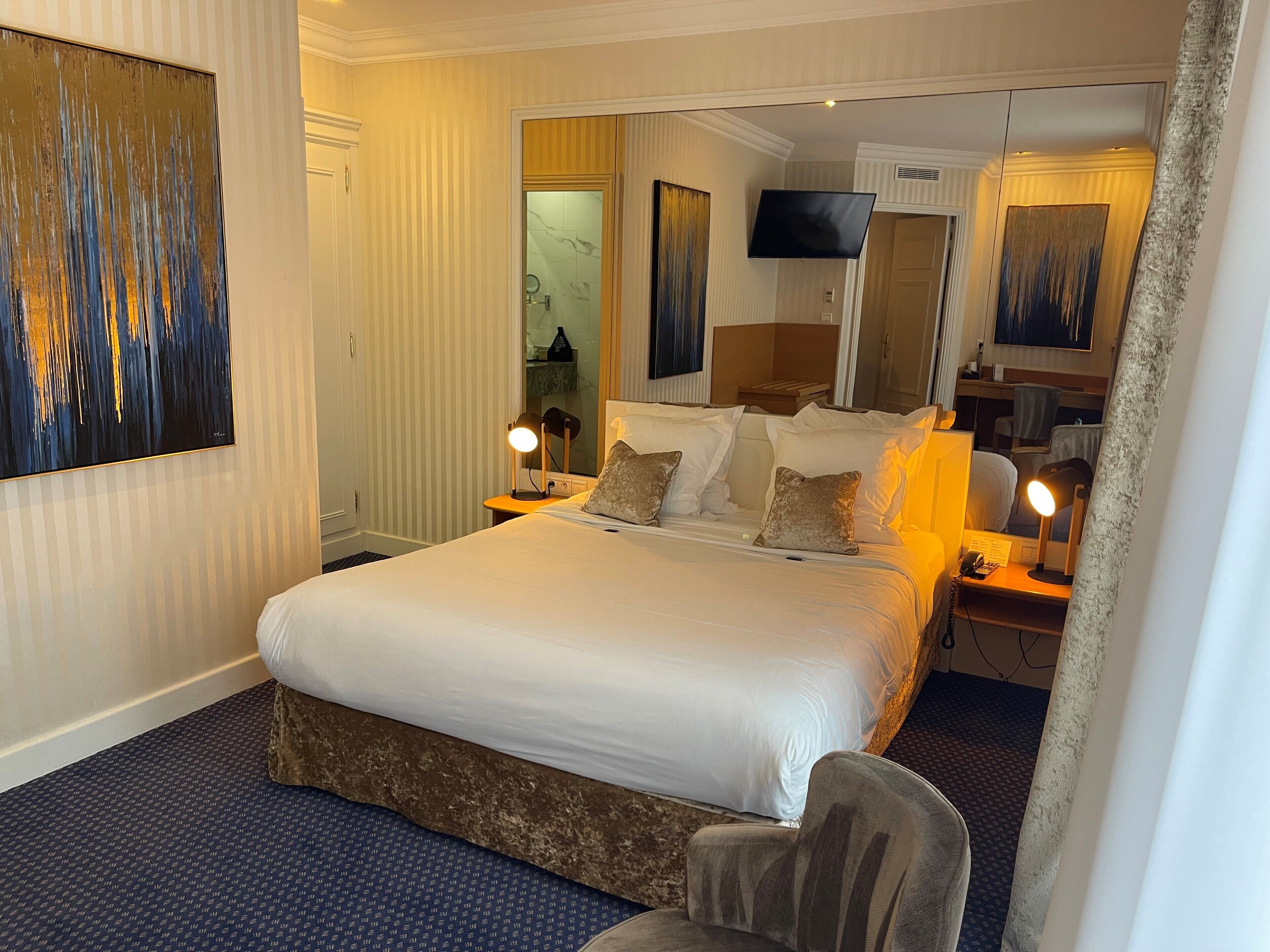 A hotel room with a large bed, two bedside tables with lamps, a flat-screen TV mounted on the wall, and a large abstract painting on the wall.