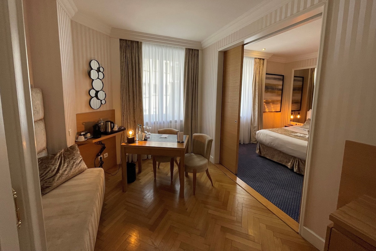 A hotel room with a bed, a desk with a chair, a coffee machine, and a small table with a lamp.