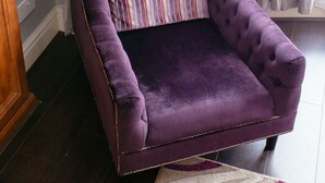 A purple velvet armchair with a striped pillow, positioned in front of a window with sheer curtains.