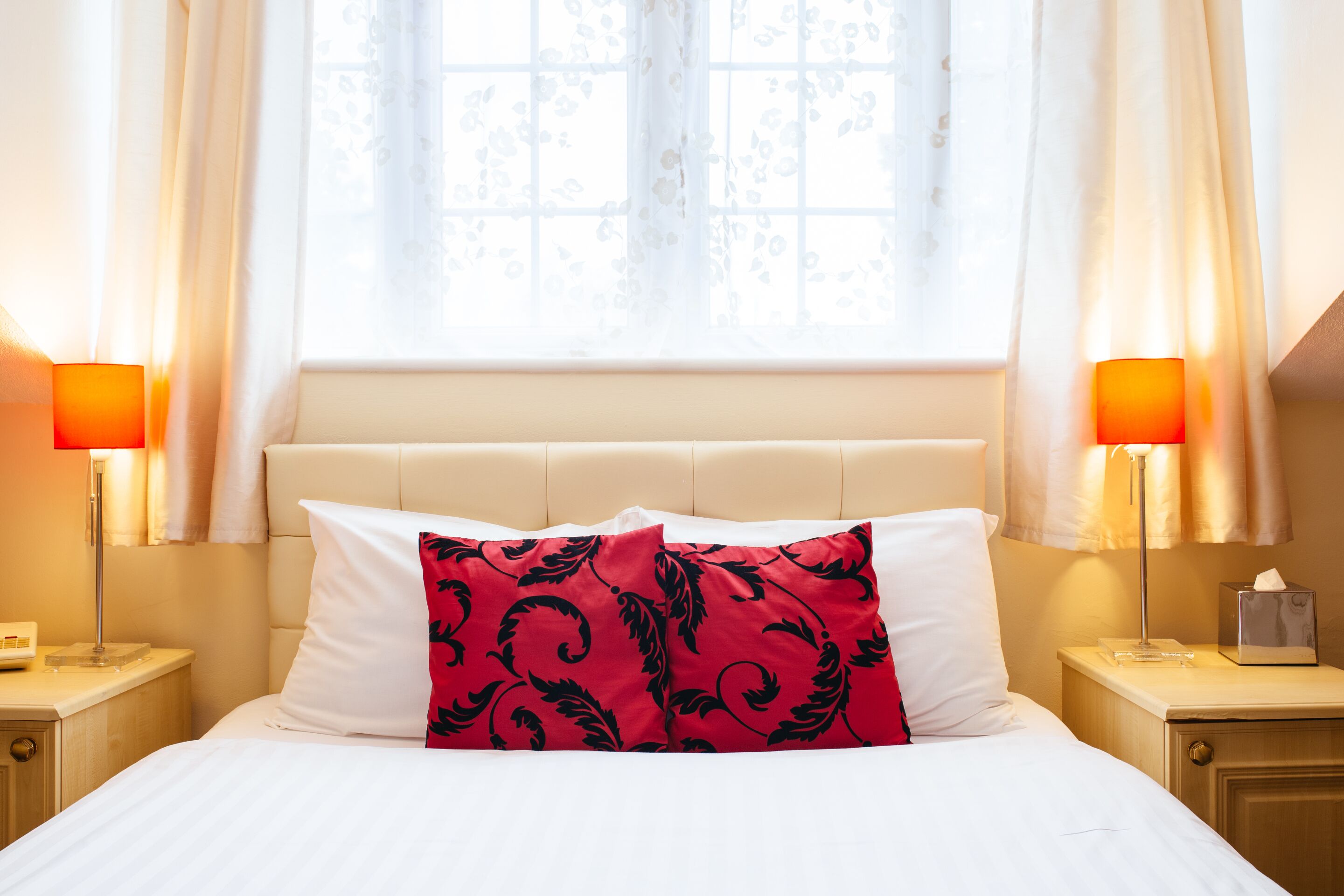 Une chambre d’hôtel avec un lit, deux lampes de chevet, une fenêtre avec des rideaux et une table de chevet sur laquelle se trouve une boîte de mouchoirs.