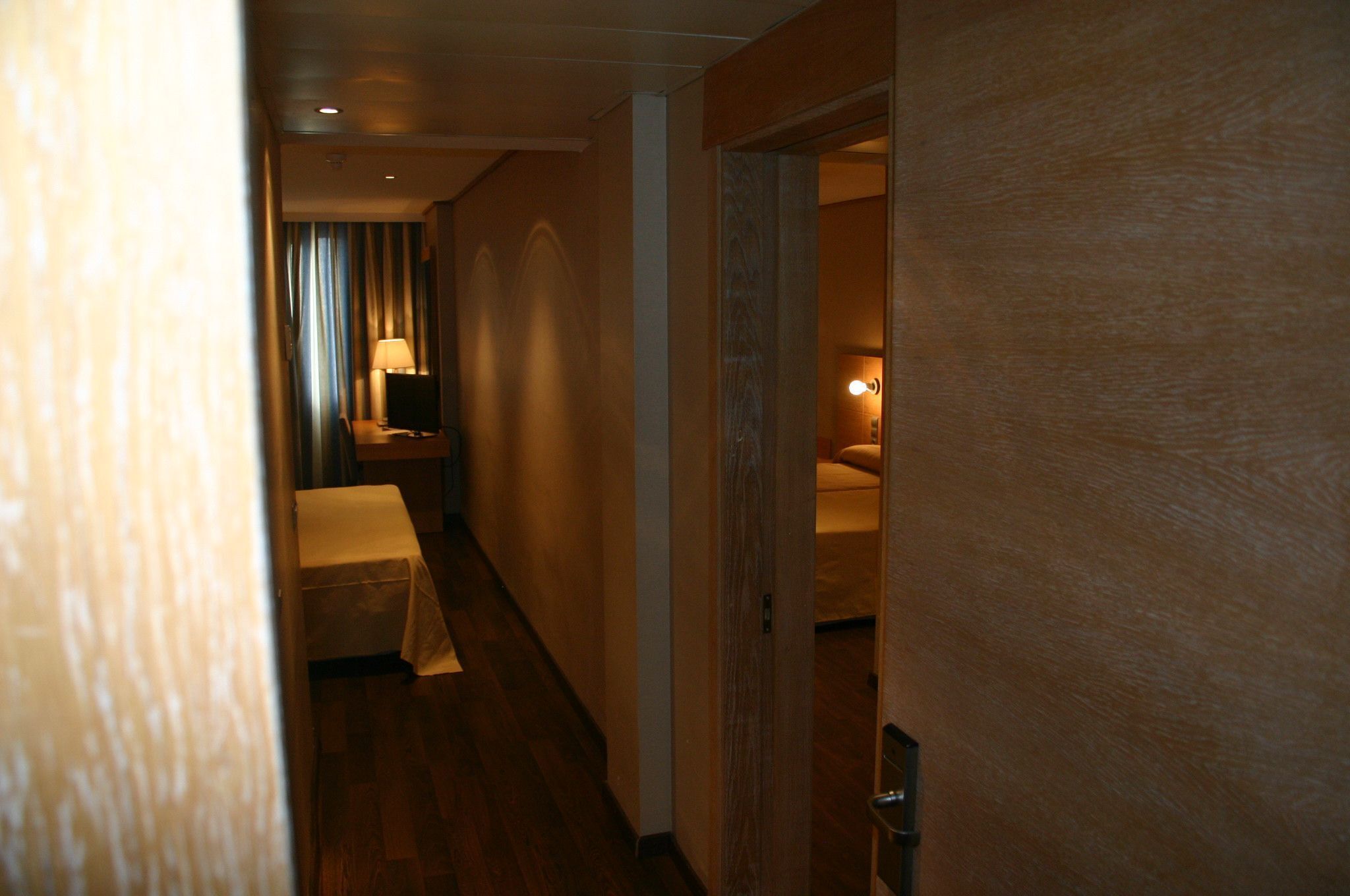 Un estrecho pasillo de hotel con suelo de madera, una cama con cubrecama blanco, una mesita de noche con lámpara y una puerta que da a otra habitación.