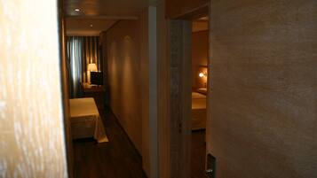 Un estrecho pasillo de hotel con suelo de madera, una cama con cubrecama blanco, una mesita de noche con lámpara y una puerta que da a otra habitación.