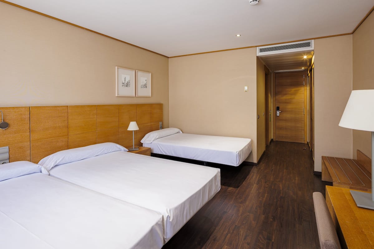 Ein Hotelzimmer mit zwei Einzelbetten, einem Holzkopfteil, einer Bank, einem Schreibtisch und einer Tür zu einem Flur.