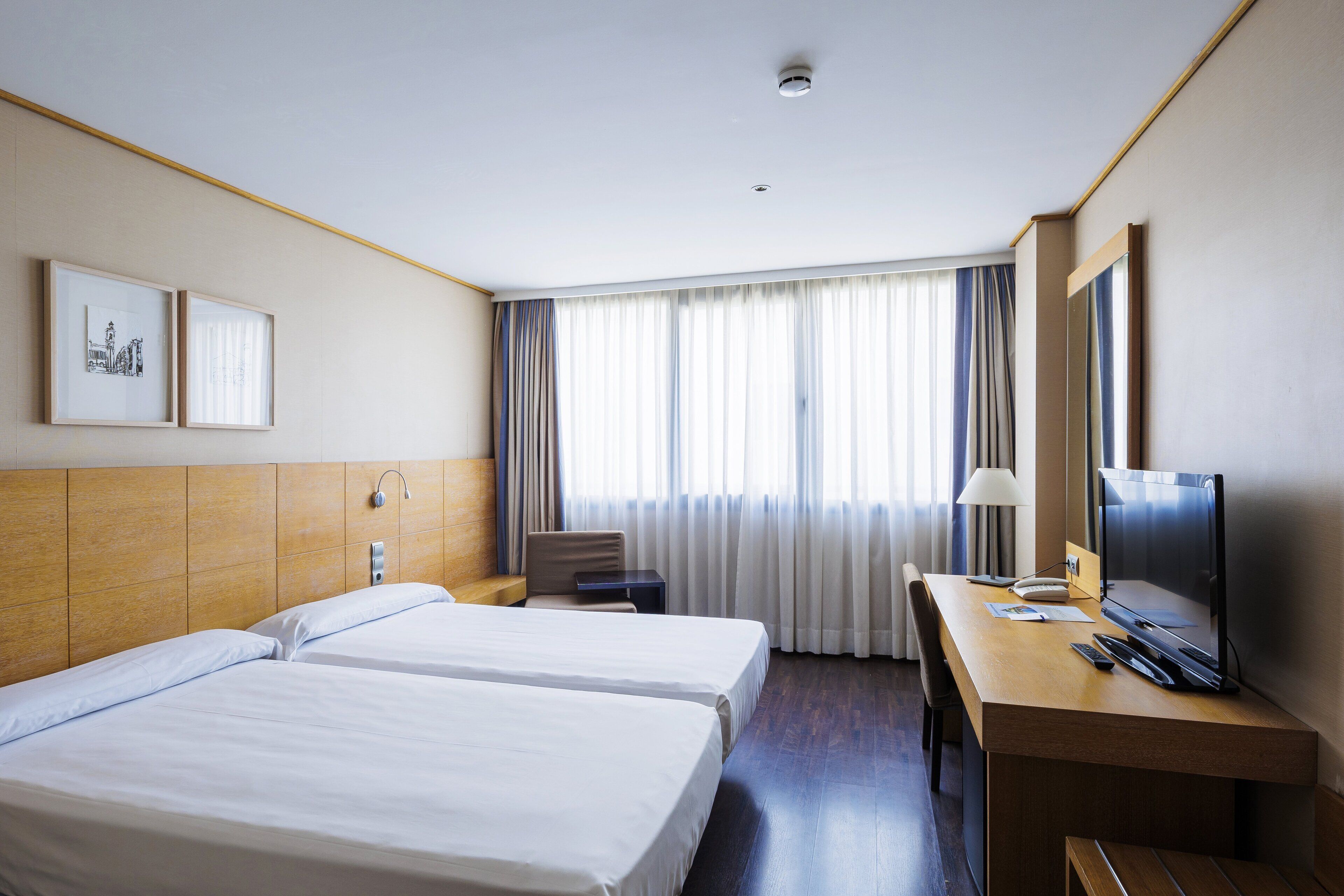 A hotel room with two beds, a desk with a TV, a chair, and a window with curtains.