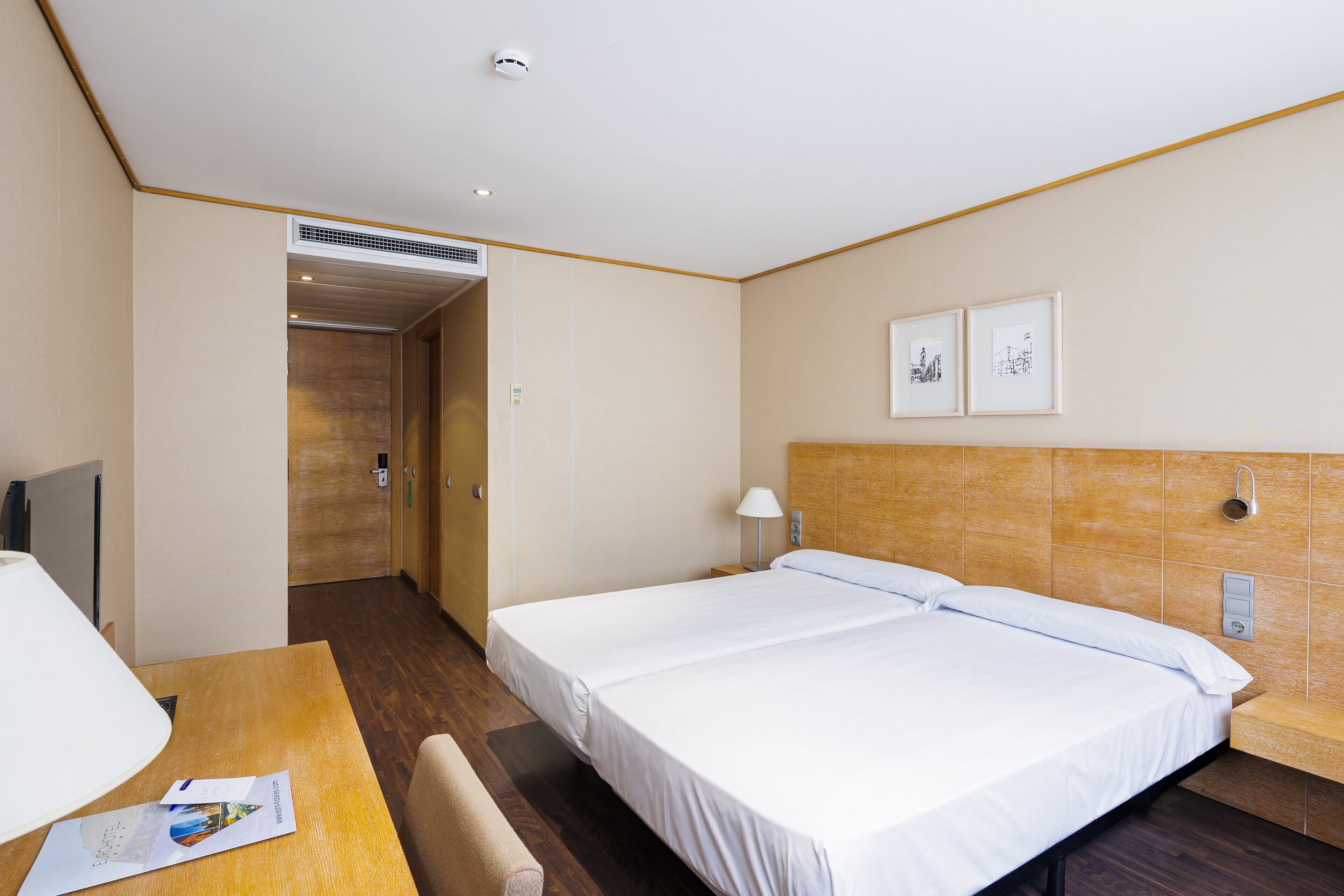 A hotel room with two beds, a desk, a lamp, and a wall-mounted TV.