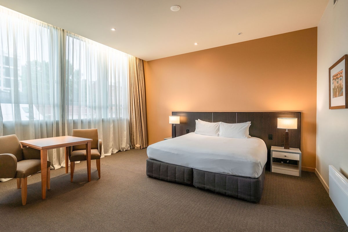 A hotel room with a large bed, a desk, two chairs, a nightstand, and a window with curtains.