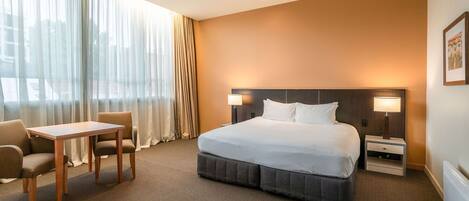 A hotel room with a large bed, a desk, two chairs, a nightstand, and a window with curtains.